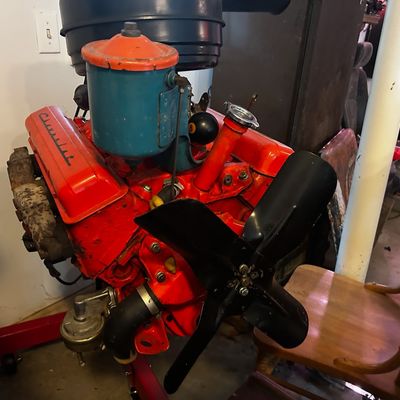 Sale Photo Thumbnail #108: Vintage Chevrolet V8 engine block mounted on a red rolling engine stand, featuring orange-painted valve covers, an oil bath air cleaner, and a black cooling fan blade.