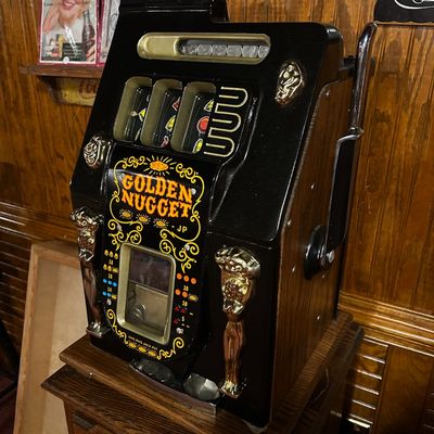 Sale Photo Thumbnail #126: Vintage Golden Nugget 25-cent mechanical slot machine with black finish, ornate gold scrollwork, and side pull handle.