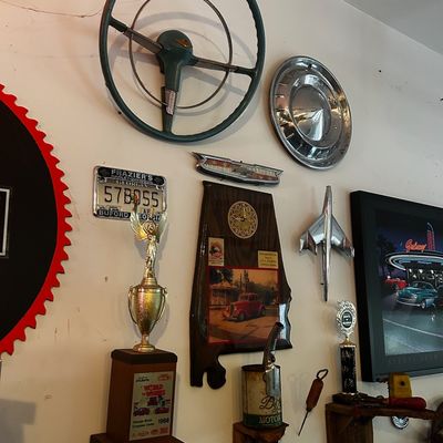 Sale Photo Thumbnail #91: Collection of vintage automotive memorabilia wall decor, including a vintage steering wheel, chrome hubcap, car emblems, license plate, and trophies.