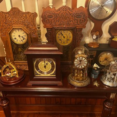 Sale Photo Thumbnail #136: Collection of antique and vintage clocks, including ornate pressed oak mantle clocks, anniversary dome clocks, and a wall-mounted barometer, displayed on a wooden console table.