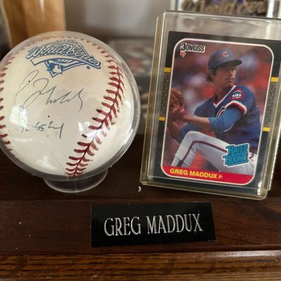 Sale Photo Thumbnail #65: Greg Maddux autographed 1995 World Series baseball alongside a 1987 Donruss Rated Rookie trading card, presented on a wooden display stand.
