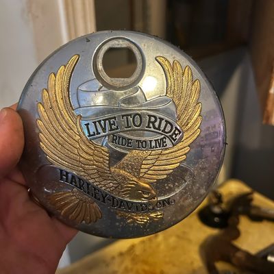 Sale Photo Thumbnail #111: Harley-Davidson "Live to Ride, Ride to Live" round chrome derby cover with gold eagle medallion detail, showing light surface patina.