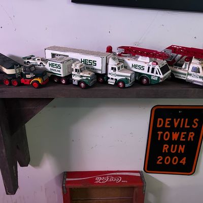 Sale Photo Thumbnail #105: Collection of vintage Hess toy trucks displayed on a wooden shelf alongside a 2004 Devils Tower Run metal sign.