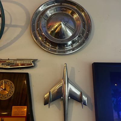 Sale Photo Thumbnail #87: Collection of vintage automotive memorabilia mounted on a wall, including a chrome Chevy hubcap with a bullet center cone and an airplane-style chrome hood ornament.