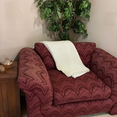 Sale Photo Thumbnail #199: Oversized burgundy flame-stitch armchair paired with a cream knitted throw blanket, set next to a wooden side table and artificial ficus tree.
