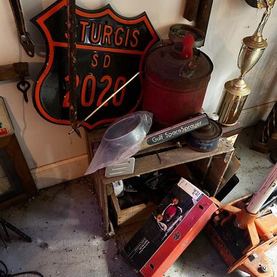 Sale Photo Thumbnail #159: Collection of garage items including a 2020 Sturgis SD metal shield sign, vintage red kerosene can, Gulf SpaceSprayer pump, trophy, and a floor jack.