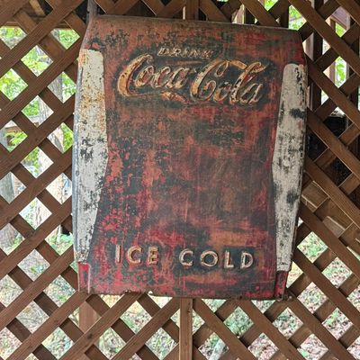 Sale Photo Thumbnail #224: Vintage painted metal Drink Coca-Cola Ice Cold sign featuring embossed lettering and significant weathering, mounted on a wooden lattice wall.