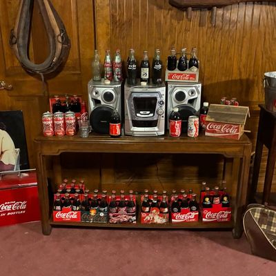 Sale Photo Thumbnail #187: Collection of Coca-Cola collectibles including vintage glass bottles in six-pack carriers, tin signs, a metal cooler, and an ice bucket set on wooden furniture alongside a Panasonic stereo system.