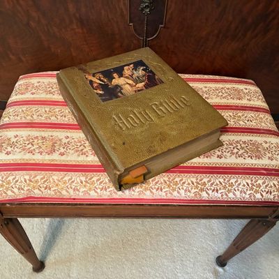 Sale Photo Thumbnail #222: Vintage illustrated Holy Bible with embossed gold-tone cover resting on a wooden bench upholstered in red and cream striped fabric.