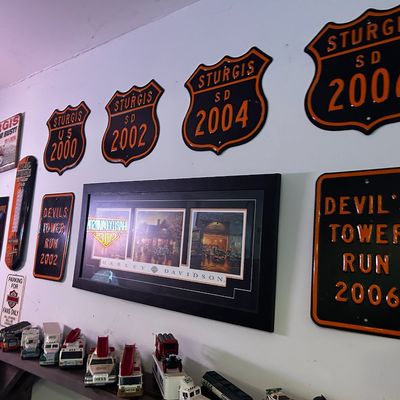 Sale Photo Thumbnail #168: A wall display of motorcycle memorabilia including Sturgis metal signs from 2000–2006, a framed Harley-Davidson print, and a collection of vintage Hess toy trucks on the shelf below.