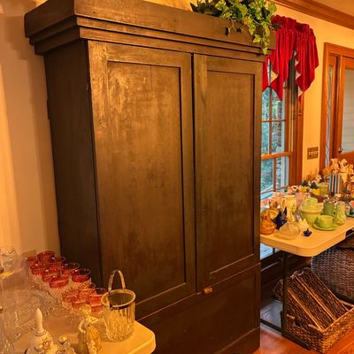 Sale Photo Thumbnail #181: Large dark-painted rustic wooden armoire or cabinet, flanked by folding tables displaying glassware, collectibles, and home decor items.