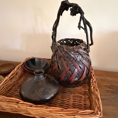 Sale Photo Thumbnail #11: Collection of Asian decorative objects including a woven bamboo ikebana basket with a rootwood handle, a small dark bronze vase featuring engraved cranes, and a matching bowl, all displayed in a woven tray.
