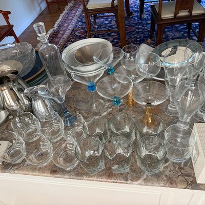Sale Photo Thumbnail #6: Large assortment of glassware and tableware, including martini glasses with colored stems, clear rocks glasses, stemware, decanters, and silver-plated serving pieces arranged on a countertop.