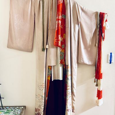 Sale Photo Thumbnail #80: Traditional Japanese silk kimono ensemble displayed on a hanging rod, featuring a pale champagne outer robe with subtle floral embroidery along the hem, layered with bright red patterned obi sashes and cords.