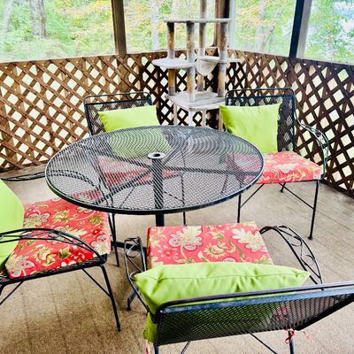 Sale Photo Thumbnail #24: Black wrought iron outdoor patio set including a round mesh table and four matching armchairs, outfitted with red floral seat cushions and bright green accent pillows.