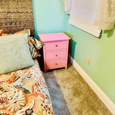 Sale Photo Thumbnail #75: Pink three-drawer nightstand set in a bedroom corner beside a bed with a woven seagrass headboard.