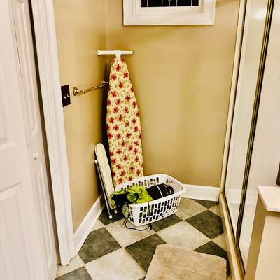 Sale Photo Thumbnail #99: A wall-mounted ironing board with floral cover, small tabletop ironing board, and white plastic laundry basket in a bathroom corner.