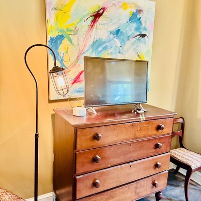 Sale Photo Thumbnail #54: Antique wooden four-drawer dresser with turned feet, topped with a flat-screen TV and framed by a large colorful abstract painting and an industrial-style gooseneck floor lamp.