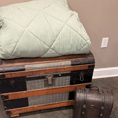 Sale Photo Thumbnail #42: Antique steamer trunk topped with a folded light green comforter, displayed alongside a smaller leather-style decorative chest.