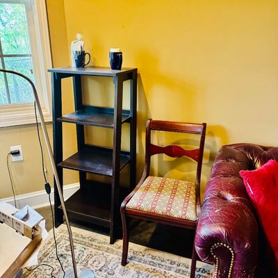 Sale Photo Thumbnail #56: Dark wood four-tier shelving unit, a vintage wooden side chair with patterned fabric seat, and a portion of a distressed red leather tufted sofa.