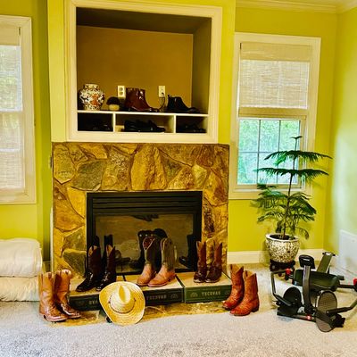Sale Photo Thumbnail #87: Collection of various men's leather cowboy boots and Tecovas shoe boxes displayed near a stone fireplace, accompanied by a straw hat, potted Norfolk Island pine, and a rowing machine.