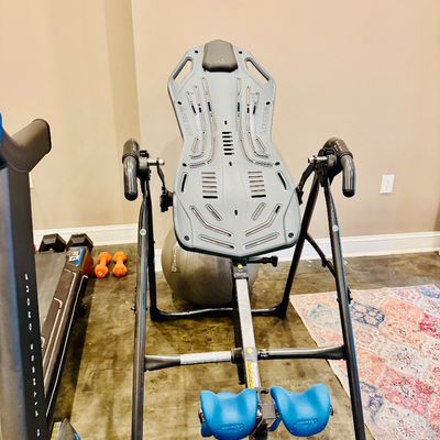 Sale Photo Thumbnail #34: Teeter Hang Ups inversion table with blue ankle supports and a gray molded backrest, shown in clean, pre-owned condition alongside other home gym equipment.