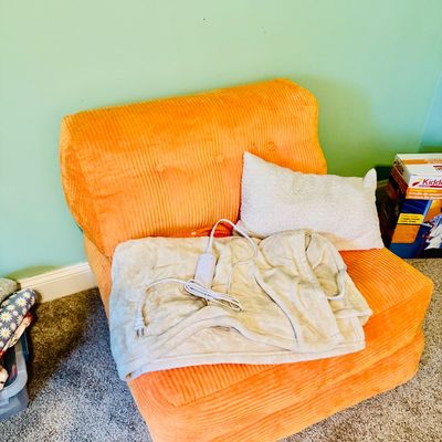 Sale Photo Thumbnail #71: Vintage armless lounge chair upholstered in bright orange corduroy, topped with a white throw pillow and a plush electric heating blanket.