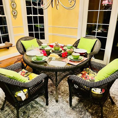 Sale Photo Thumbnail #94: Round dark wicker dining table with glass top and set of four matching woven armchairs featuring vibrant green and floral cushions, fully set with assorted tableware.