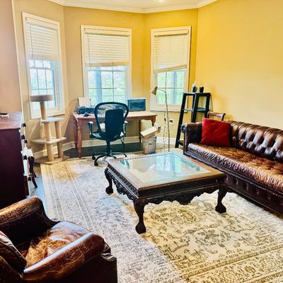 Sale Photo Thumbnail #47: Living room setup featuring a distressed brown leather chesterfield sofa, matching armchair, carved glass-top coffee table, large area rug, and office desk.