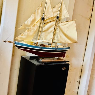 Sale Photo Thumbnail #45: Handcrafted wooden model sailboat with cream-colored cloth sails, displayed on a wooden stand above a black pedestal.