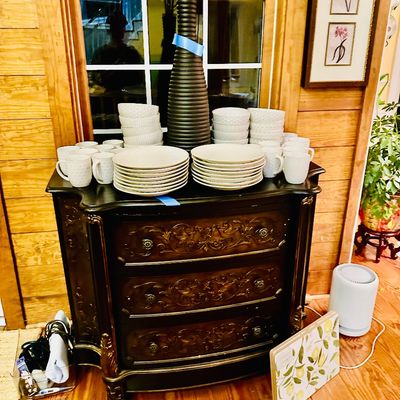 Sale Photo Thumbnail #93: Ornate French Provincial style dark wooden chest with carved three-drawer front, displayed alongside stacks of cream-colored ceramic dinnerware and a tall ribbed vase.