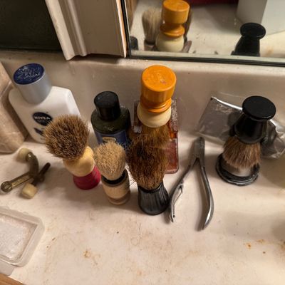 Sale Photo Thumbnail #67: Collection of vintage shaving brushes with various wooden and plastic handles alongside grooming tools on a bathroom counter.