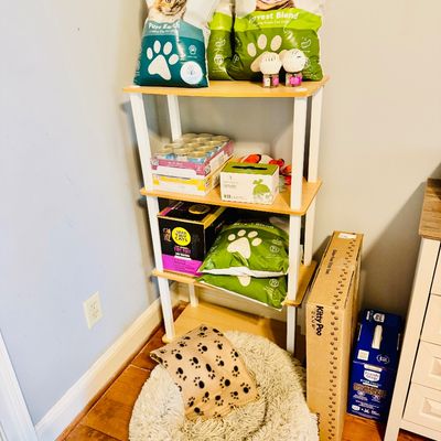 A light wood and white frame four-tier shelving unit loaded with various cat supplies, including bags of Kitty Poo Club litter, canned...