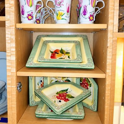 Sale Photo Thumbnail #94: Collection of floral botanical ceramic mugs and fruit-motif square serving platters displayed inside a light wood cabinet.