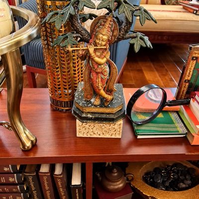Sale Photo Thumbnail #76: Intricately detailed Hindu deity statue of Krishna playing a flute under a tree, mounted on a stone pedestal atop a wooden shelf.