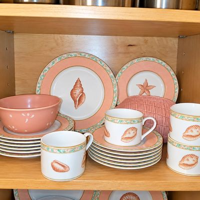 Sale Photo Thumbnail #103: Set of coastal-themed dishware featuring seashell motifs, peachy pink borders, matching teacups, saucers, and plates arranged in a cabinet.