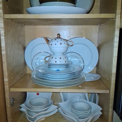 Sale Photo Thumbnail #105: A light wood cabinet filled with various china service pieces, including a star-patterned teapot and teacup set, dinner plates, and decorative bowls.