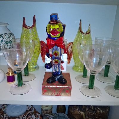 Sale Photo Thumbnail #111: Murano-style Venetian glass clown decanter standing among green-stemmed wine glasses, art glass vases, and marble decorative items.