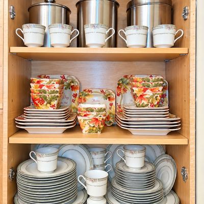 Sale Photo Thumbnail #95: Wooden cabinet filled with tableware including a fine porcelain china set, autumn-themed leaf motif dishware, and stainless steel kitchen canisters.