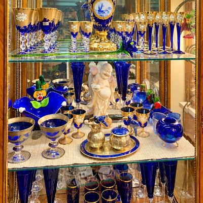 Sale Photo Thumbnail #72: Collection of cobalt blue and gold rimmed glassware displayed in a lit curio cabinet, including stemware, a large porcelain vase, and decorative figurines.