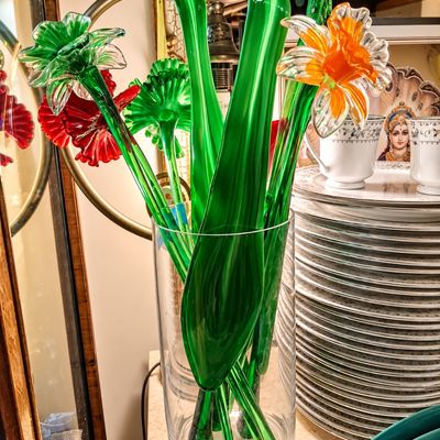 Sale Photo Thumbnail #102: Collection of vibrant handcrafted Murano-style glass flowers with long green stems, displayed in a clear cylindrical vase alongside fine china dinnerware.