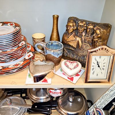Sale Photo Thumbnail #92: Collection of decorative collectibles and kitchenware on a shelf, including Imari-style china plates, a bronze-tone Mount Rushmore plaque, a quartz desk clock, heart-shaped trinket boxes, and various stainless steel cookware below.