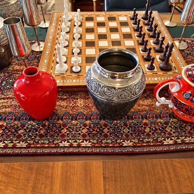 Sale Photo Thumbnail #74: Inlaid wooden chess set displayed alongside a collection of silver-plated goblets, embossed metallic urns, and colorful ceramic vessels on a pattern table runner.