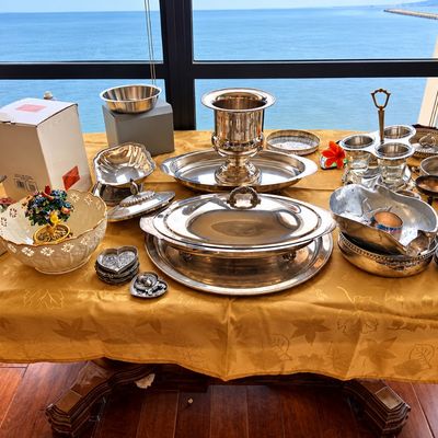 Sale Photo Thumbnail #77: Collection of silver-plated serveware including a wine cooler, covered chafing dish, trays, and condiment caddy displayed on a table alongside decorative ceramic bowls.