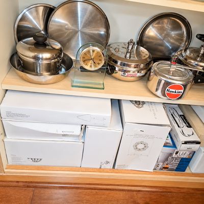 Sale Photo Thumbnail #100: Shelves filled with stainless steel cookware including Hawkins and Vinod pressure cookers, along with a glass desk clock and boxed home goods.