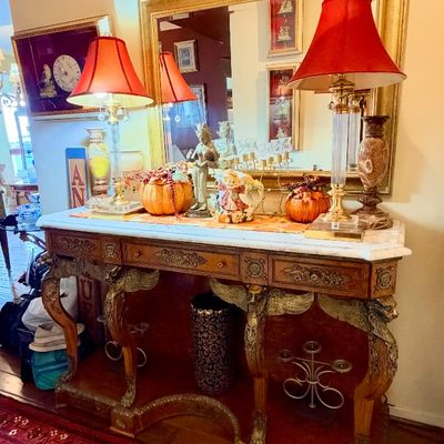 Sale Photo Thumbnail #83: Ornate French Empire style marble-top console table featuring carved winged griffin legs, paired with a matching gilt framed wall mirror and brass table lamps with red fabric shades.