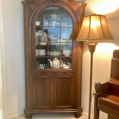 Traditional dark wood corner hutch with shell motif cresting, arched glass door, and bottom cabinet storage.



