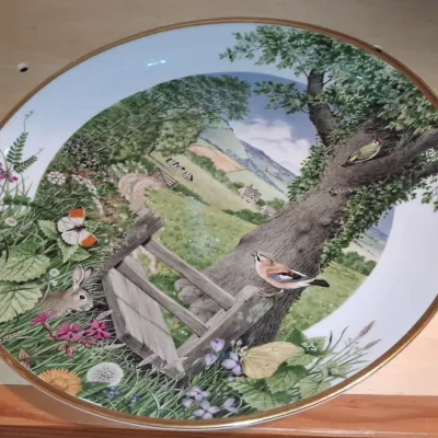 Sale Photo Thumbnail #673: Decorative porcelain collector plate featuring an illustrated countryside scene with birds, a rabbit, and wildflowers, trimmed with a gold-toned rim.