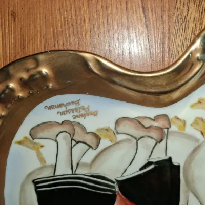 Sale Photo Thumbnail #600: Detail view of a hand-painted ceramic tray featuring figures in traditional Dutch attire and an artist signature along the gilded rim.