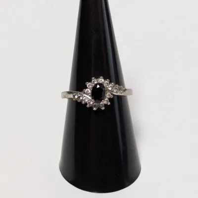 Sale Photo Thumbnail #795: Silver-tone ring featuring a central oval dark gemstone surrounded by a halo and bypass band of clear accent stones, presented on a black cone display.